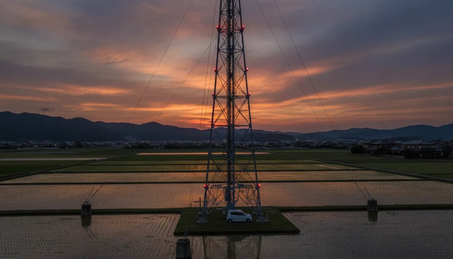夕暮れの水田地帯に立つ、支線で支えられた鉄塔。塔は画面の上端を超えて伸び、赤い航空障害灯が点いている。根元には軽自動車が1台停まっていて、その対比から塔がきわめて高いことが分かる。