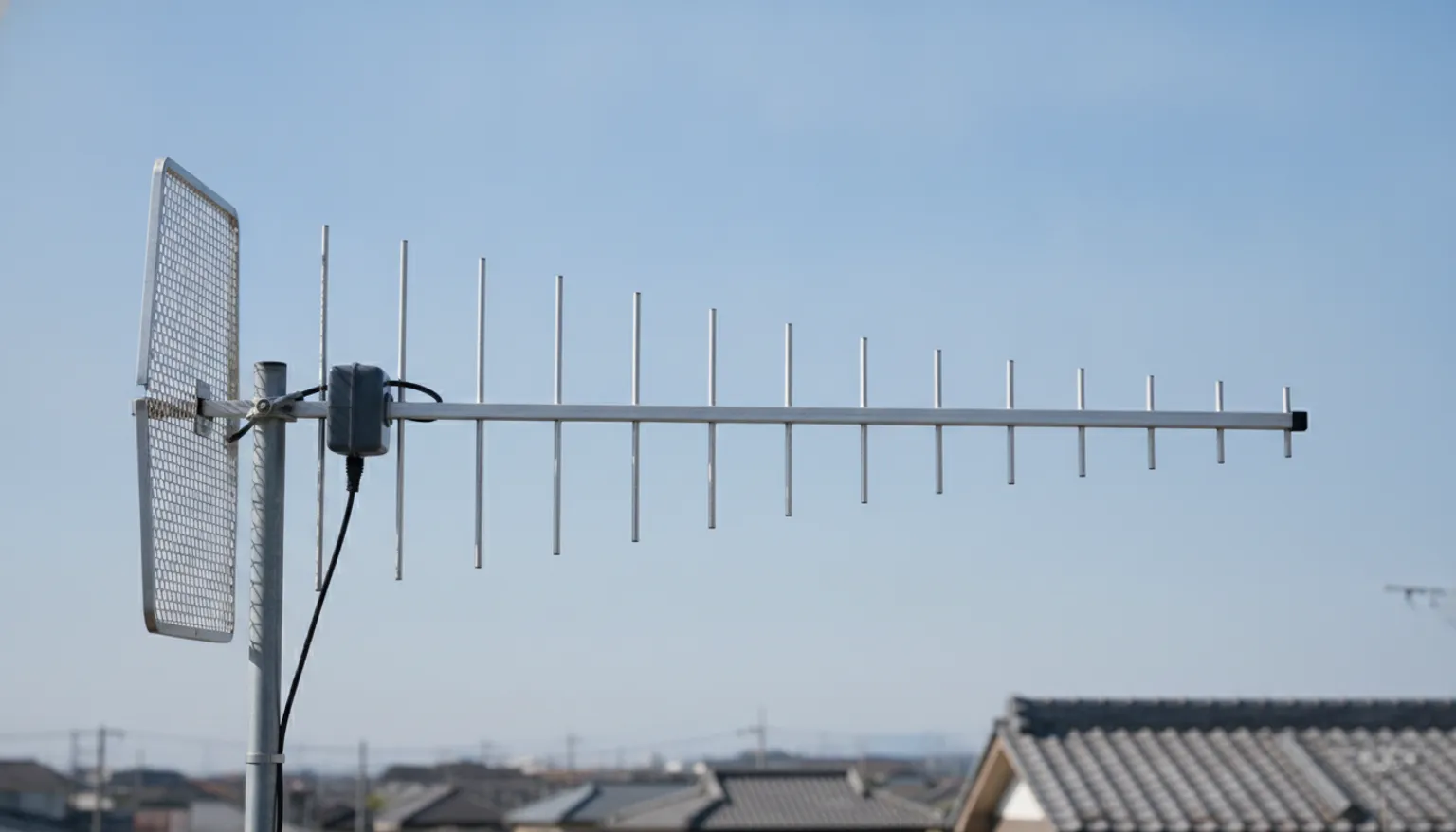 青空を背景に横から見た八木・宇田アンテナ。左端に格子状の反射器と、給電箱と同軸ケーブルの付いた輻射器があり、そこから右へ向かって十数本の素子が少しずつ短くなりながら並んでいる。背景には日本の瓦屋根の住宅が写っている。